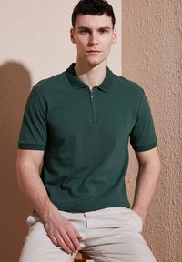 Tamsiai žalia zip-up polo marškinėliai su trumpomis rankovėmis, tekstūruota medžiaga ir kontrastinga gumbinė apykaklė, derinama su šviesios spalvos kelnėmis.