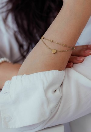 Gold double-chain bracelet with small beads and a heart charm worn on a raised forearm, person in white long-sleeve shirt.