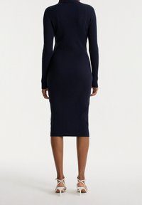 Robe midi côtelée bleu marine à manches longues et col haut. Portée avec des sandales à talons hauts blanches à lanières, elle présente une silhouette ajustée.