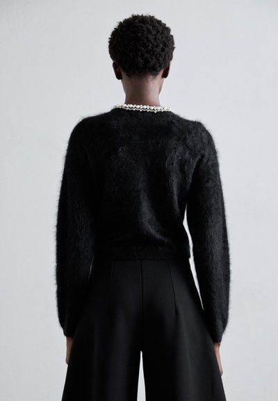 Schwarzer, flauschiger Pullover mit langen Ärmeln, tailliert. Kombiniert mit schwarzen, weit geschnittenen Hosen. Eine Perlenkette akzentuiert den Ausschnitt.