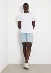 Camiseta de algodón blanca combinada con shorts de mezclilla azul claro, doblados en el dobladillo, completados con zapatillas de deporte en color blanco roto sobre un fondo neutro.