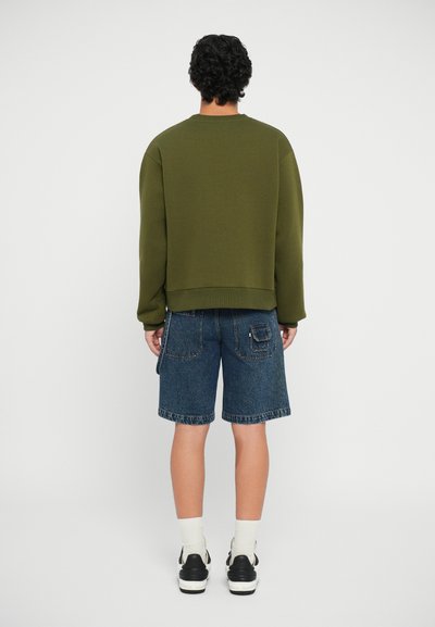 Felpa verde oliva con una vestibilità ampia, abbinata a pantaloni corti di denim fino al ginocchio. Calzini bianchi e sneakers nere completano l'outfit.