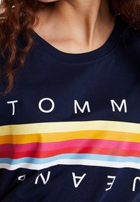 Camiseta azul marino con cuello redondo que presenta en letras blancas gruesas la palabra "TOMMY" y franjas horizontales superpuestas en rojo, rosa, amarillo y azul.