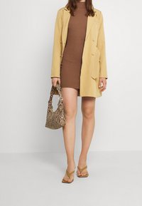 Kvinde iført en brun ribbet kjole og en beige jakke, holder en taske med slangeskindsprint og bærer beige højhælede sandaler.