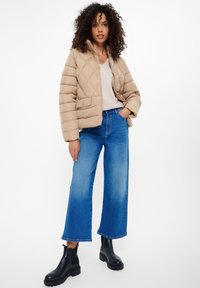 Beżowa pikowana kurtka puchowa z wysokim kołnierzem, w połączeniu z jasnoniebieskimi dżinsami o szerokich nogawkach i czarnymi botkami za kostkę.
