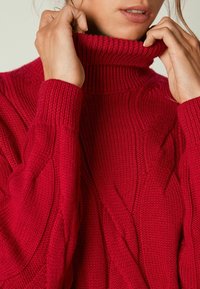 Femme ajustant le col montant d'un pull rouge en maille torsadée, montrant la texture détaillée et les motifs de point.