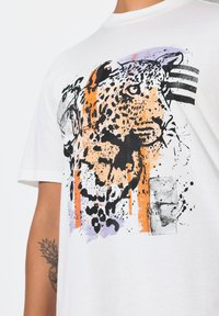 Bílý bavlněný tričko s leopardím grafickým potiskem v černé, oranžové a fialové barvě s detaily ve stylu rozstřiku. Krátký rukáv a design s kulatým výstřihem.