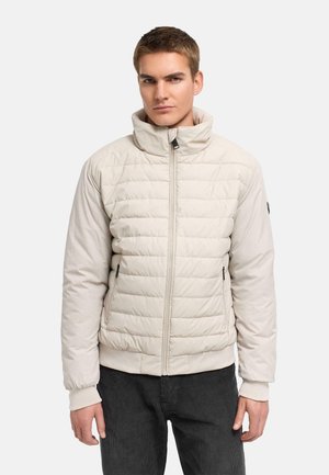 Ung mand iført en lys beige quiltet jakke med høj krave og sorte bukser, vendt fremad mod en ensfarvet baggrund.