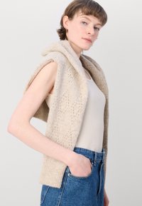 Gilet beige in maglia con cappuccio, caratterizzato da un motivo a pizzo e bordi a coste. Indossato sopra una canotta leggera e abbinato a jeans di denim.