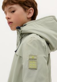 Lichtgroene waterdichte jas met een grote kap, zijzakken en een getextureerde patch met een schedellogo op de mouw.
