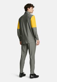 Atletický teplákový komplet v sivej farbe s žltými ramenami. Obsahuje vysoký golier a slim-fit nohavice, navrhnuté pre pohodlie a pohyb.