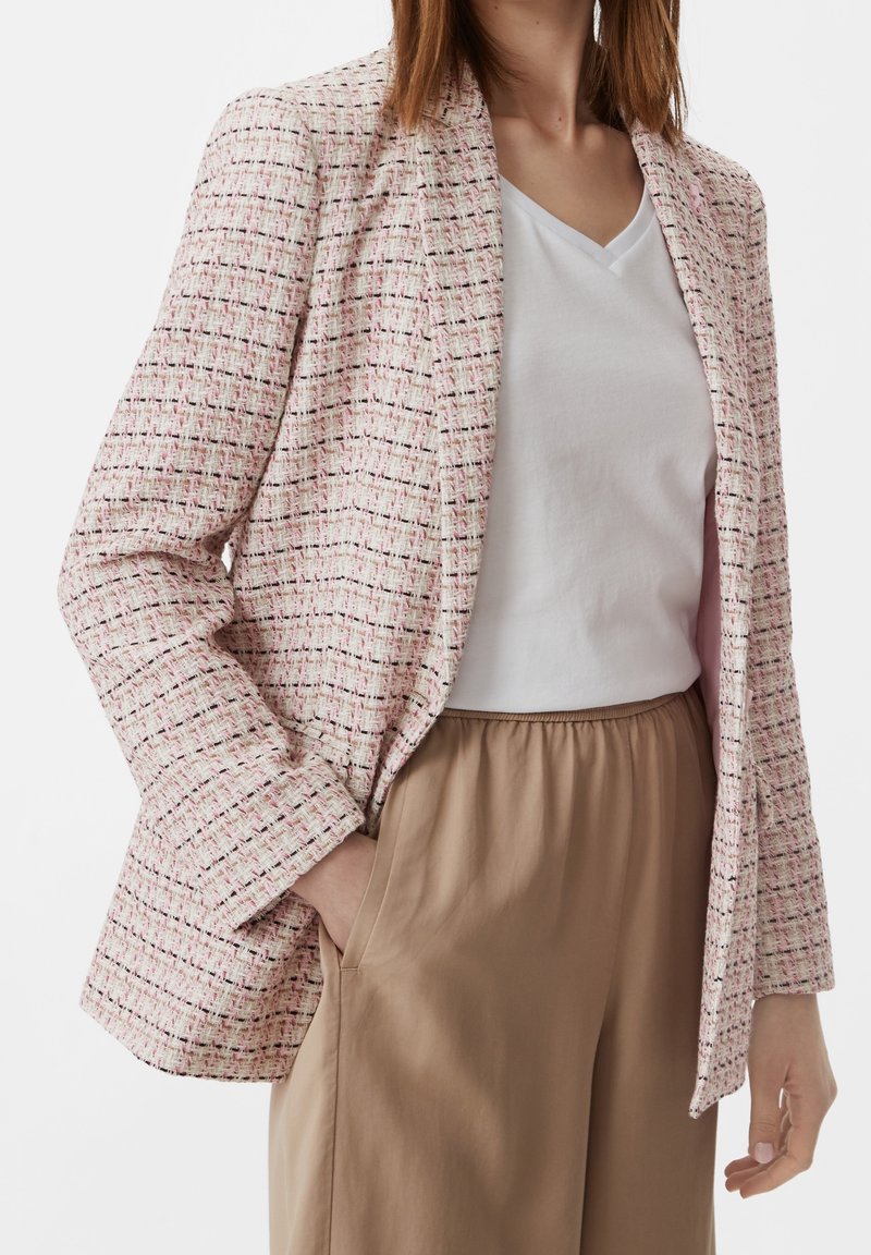 Femme portant un blazer en tweed rose clair, un t-shirt blanc à col en V et un pantalon beige, une main dans la poche, face à l'avant.