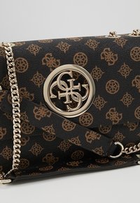 Sac à main noir à motif avec un logo circulaire doré, une bandoulière chaîne, et une finition en cuir texturé avec des accents marron.