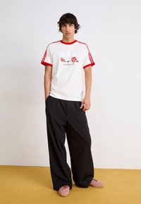 Valkoinen t-paita, jossa on punainen kaulus ja hihat, ja jossa on kukkakuva ja teksti. Mustat, löysät housut ja vaaleanpunaiset kengät.