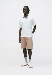 Lys blå kortermet teksturert skjorte, beige teksturerte shorts, hvite sko og grå sokker, modellert av en person i en nøytral setting.