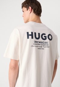 Cremefarbendes Baumwoll-T-Shirt mit großem navyblauen "HUGO"-Logo und Schriftzug auf der Rückseite, kurzen Ärmeln und lockerer Passform.