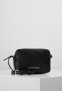 Borsa in pelle nera con design in rilievo "NY" e testo "Calvin Klein". Forma rettangolare, tracolla regolabile e chiusura con zip. Tessuto liscio.