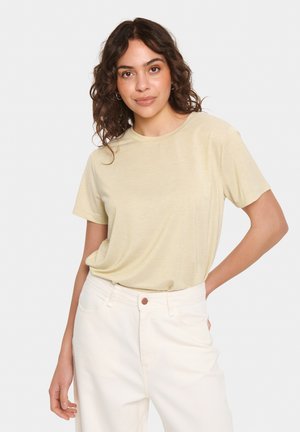 T-shirt à manches courtes, col rond en jaune clair avec un subtil éclat, associé à un pantalon blanc taille haute avec un bouton marron.