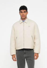 Šviesiai smėlinis pūkinis švarkas su priekiniais užtrauktukais, dideliu apykaklės, ir dviem šoniniais kišenėmis. Audinys atrodo lygus su šiek tiek blizgiu paviršiumi.