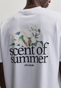 Uomo che indossa una maglietta bianca con fiori bianchi e foglie verdi e la scritta "profumo d'estate" sulla schiena.