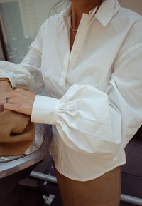 Witte katoenen shirt met poffende mouwen en knoopmanchetten, gedragen met een bruine stof gedrapeerd in de handen. Eenvoudig ontwerp, gladde textuur.