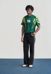 Πράσινο αθλητικό πουκάμισο με κίτρινες λεπτομέρειες και αριθμό 94, συνδυασμένο με μαύρο τζιν φαρδιάς γραμμής και άσπρα παπούτσια, που στέκεται σε μπλε χαλί.