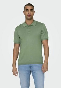 Polo shirt in cotone verde con colletto, maniche corte e chiusura a tre bottoni; indossato con jeans azzurri chiari.