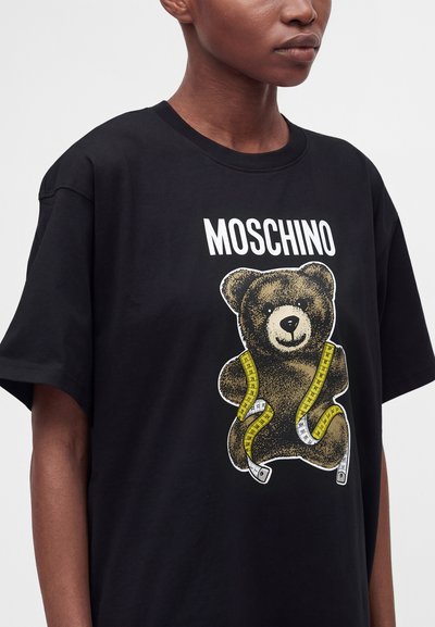 Άτομο που φοράει μαύρο T-shirt με μια αρκούδα τυλιγμένη σε κίτρινη μεζούρα και τη λέξη MOSCHINO πάνω από την αρκούδα.
