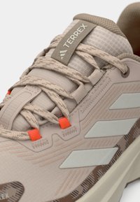 Chaussure de sport beige clair avec tige en mesh et accents texturés. Comprend des rayures grises, des œillets orange et une semelle extérieure flexible.