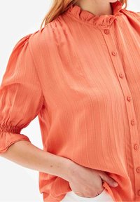 Blouse pêche avec un motif rayé texturé, fermeture boutonnée, col à volants et poignets élastiques. Tissu léger qui tombe en douceur.