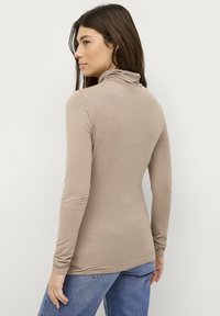 Langärmeliges, Rollkragenoberteil aus leicht beigem Stoff, das eine taillierte Silhouette und eine weiche Textur aufweist. Kombiniert mit blauen Denim-Jeans.