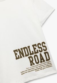 Biała bawełniana koszulka z ciemnobrązowym napisem "ENDLESS ROAD" oraz dodatkowymi frazami związanymi z przygodą w mniejszej czcionce z boku.