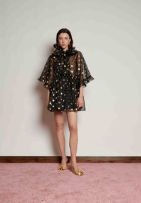 Μαύρο διάφανο φόρεμα με χρυσές πουά, φαρδιά μανίκια και βολάν στο ντεκολτέ; συνδυασμένο με τακούνια με χρυσή φιόγκους. Τοποθετημένο σε ανοιχτό φόντο.