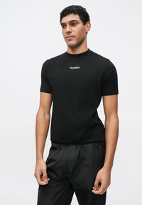 Juoda trumpomis rankovėmis marškinėliai iš minkštos medžiagos, suapvalintu iškirpte ir mažo balto logotipo centruojamu centre. Dėvimi su juodomis lengvomis kelnėmis.