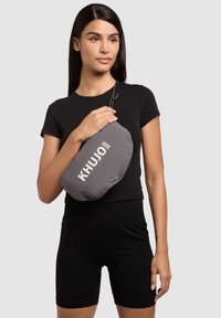 Femme en tenue noire tenant une pochette grise de la marque Khujo avec un cordon, devant un fond clair uni.