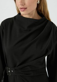 Černé šaty s vysokým výstřihem, nařasenou látkou a pásem v pase. Měkká textura s hladkým povrchem a minimalistickými designovými detaily.