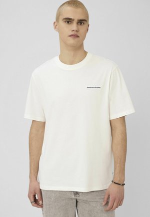 Jeune homme aux cheveux blonds courts portant un t-shirt blanc avec un petit texte sur la poitrine, un jean gris clair et un bracelet noir en perles.
