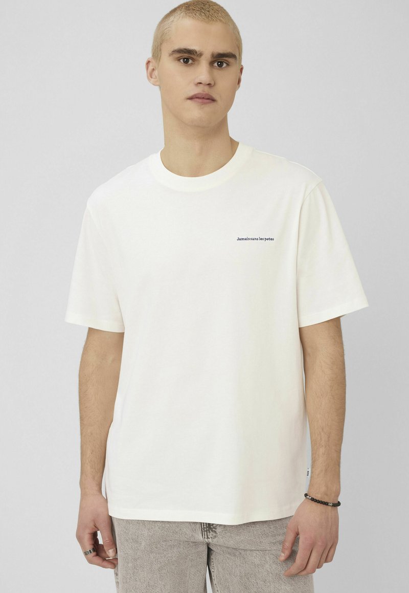 Jeune homme aux cheveux blonds courts portant un t-shirt blanc avec un petit texte sur la poitrine, un jean gris clair et un bracelet noir en perles.