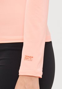 Maglietta a maniche lunghe in tessuto rosa chiaro con una texture liscia. Presenta un piccolo logo UVSURF arancione sulla manica sinistra.