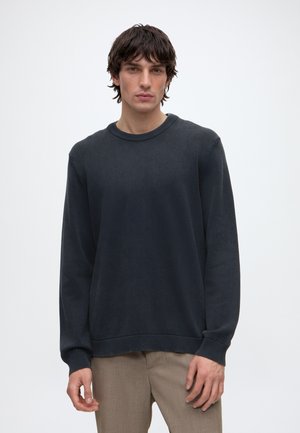 Jeune homme aux cheveux foncés porte un pull bleu marine à manches longues et un pantalon marron clair, debout devant un fond clair uni.