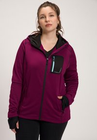 Haut à capuche fuchsia zippé avec une capuche noire, doté d'une poche à zip sur le devant avec un zip bleu, des manches longues et des poignets avec trou pour le pouce.
