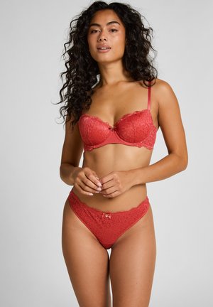 Vrouw met lang krullend haar, gekleed in bijpassende rode kanten beha en slipje, staand met de handen gevouwen voor zich tegen een effen achtergrond.