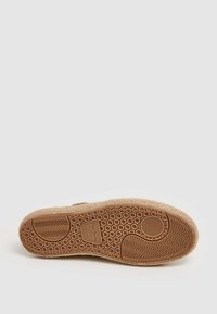 Suela de zapato con textura de panal en goma marrón claro, con un acento circular y el logotipo de la marca, que ofrece flexibilidad, agarre y durabilidad.