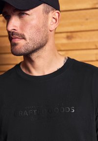 Muž s vousy a čepicí, oblečený v černém tričku s vyraženým textem "Crafted Goods", na dřevěném pozadí.