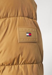 Brown puffer vest with quilting detail and a textured fabric. Features a tan branded patch with a blue, white, and red emblem.