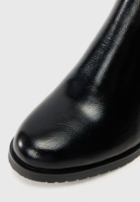 Stivale alla caviglia nero con tomaia sintetica lucida, punta arrotondata e finitura texturizzata. Presenta una suola in gomma piatta con un leggero motivo di aderenza.
