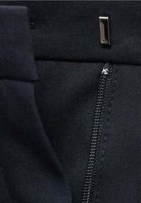 Gros plan sur un vêtement en tissu noir avec une fermeture éclair métallique partiellement ouverte et une boucle métallique rectangulaire au-dessus.