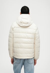 Γεμιστό μπουφάν σε εκρού, με κουκούλα, ελαστικές μανσέτες και ραμμένο σχέδιο. Φοριέται με ανοιχτό μπλε τζιν.