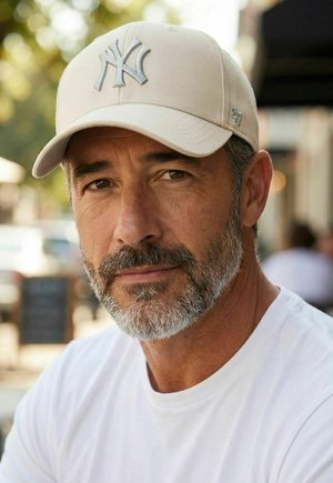 Mittelalter Mann mit salt-and-pepper Bart, der eine beige Baseballmütze und ein weißes Hemd trägt und draußen direkt in die Kamera blickt.