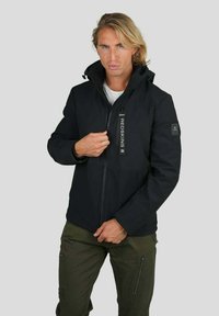 Veste noire zippée avec un col haut, finition mate, patch de marque subtile sur la manche et deux poches avant. Associée à un pantalon olivier.